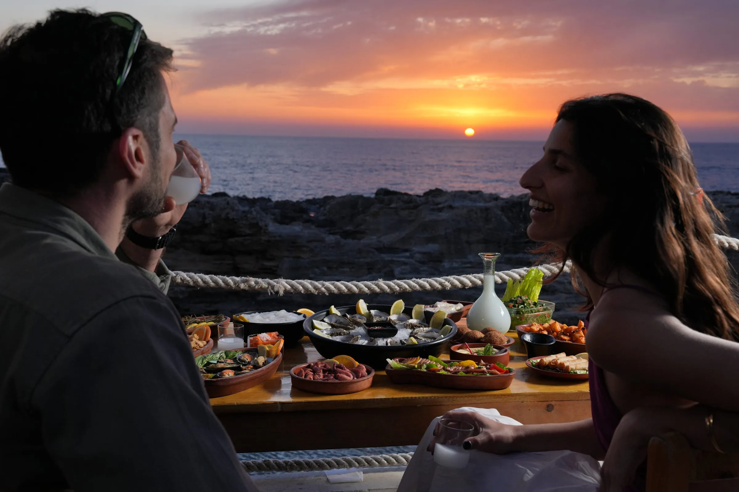 Two guests at a seafood table by the sea, watching the sun go down