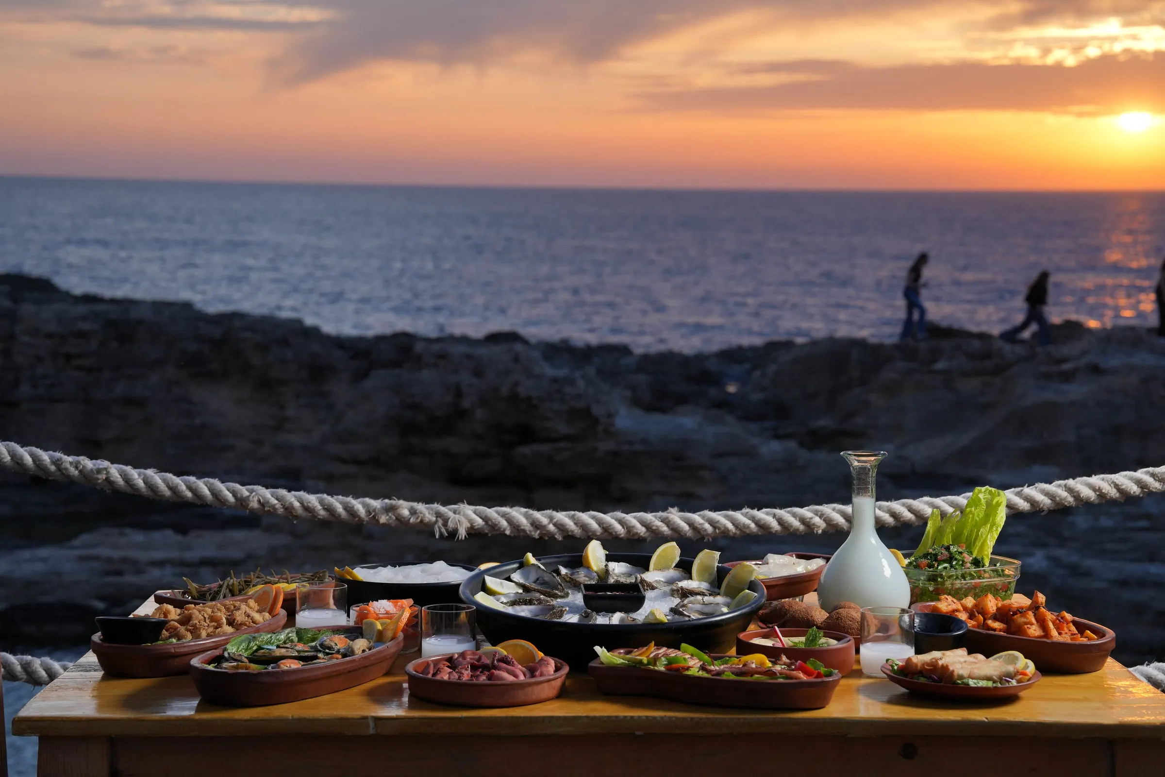 A table of seafood set by the sea at sunset, two people walking on the rocks behind