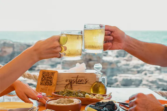 Two glasses of beer clinking over a table by the sea at Oyster's Seafood Bar, Batroun